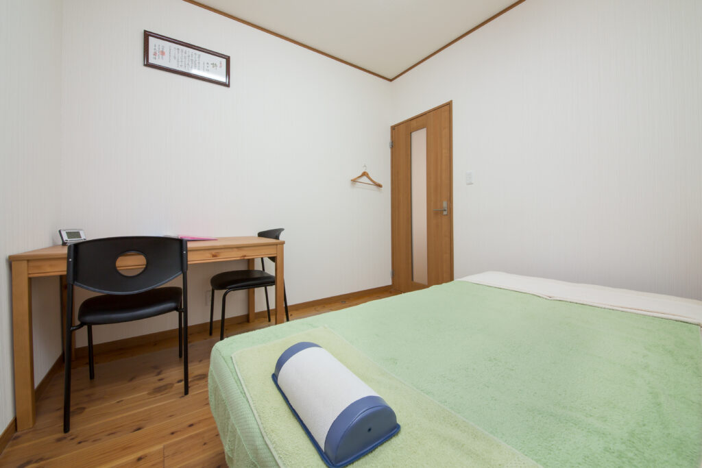 個室の施術室。緑系シーツの施術台と足元のクッション、白壁と木床、壁側に木製デスクと椅子が並ぶ清潔な院内|山下針灸房